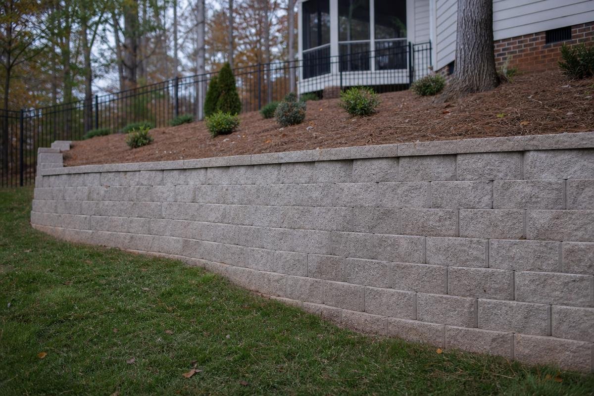 Tall gray segmental block retaining wall along a sloped residential property in Waxhaw, NC, with a mulched hillside and evergreen shrubs above
