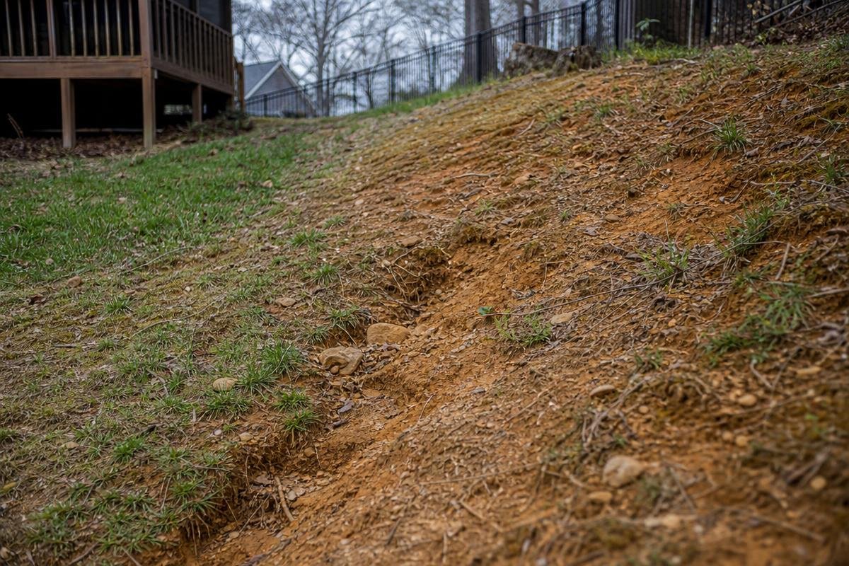 Eroded red clay hillside with runoff channels and sparse grass on a sloped residential backyard in Waxhaw, NC, showing the need for a retaining wall